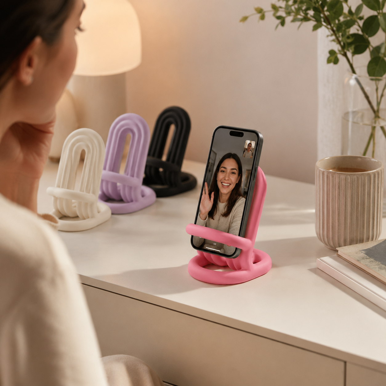 A pink phone stand holding a smartphone in a video call, shown on a white desk in a warm indoor setting. Three additional phone stands in white, lavender, and black are visible in the background, along with a lamp, plant, and cup.