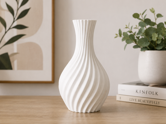A white ribbed ceramic vase with a flared neck and bulbous base, displayed on a light wood tabletop next to a potted plant and a stack of books.