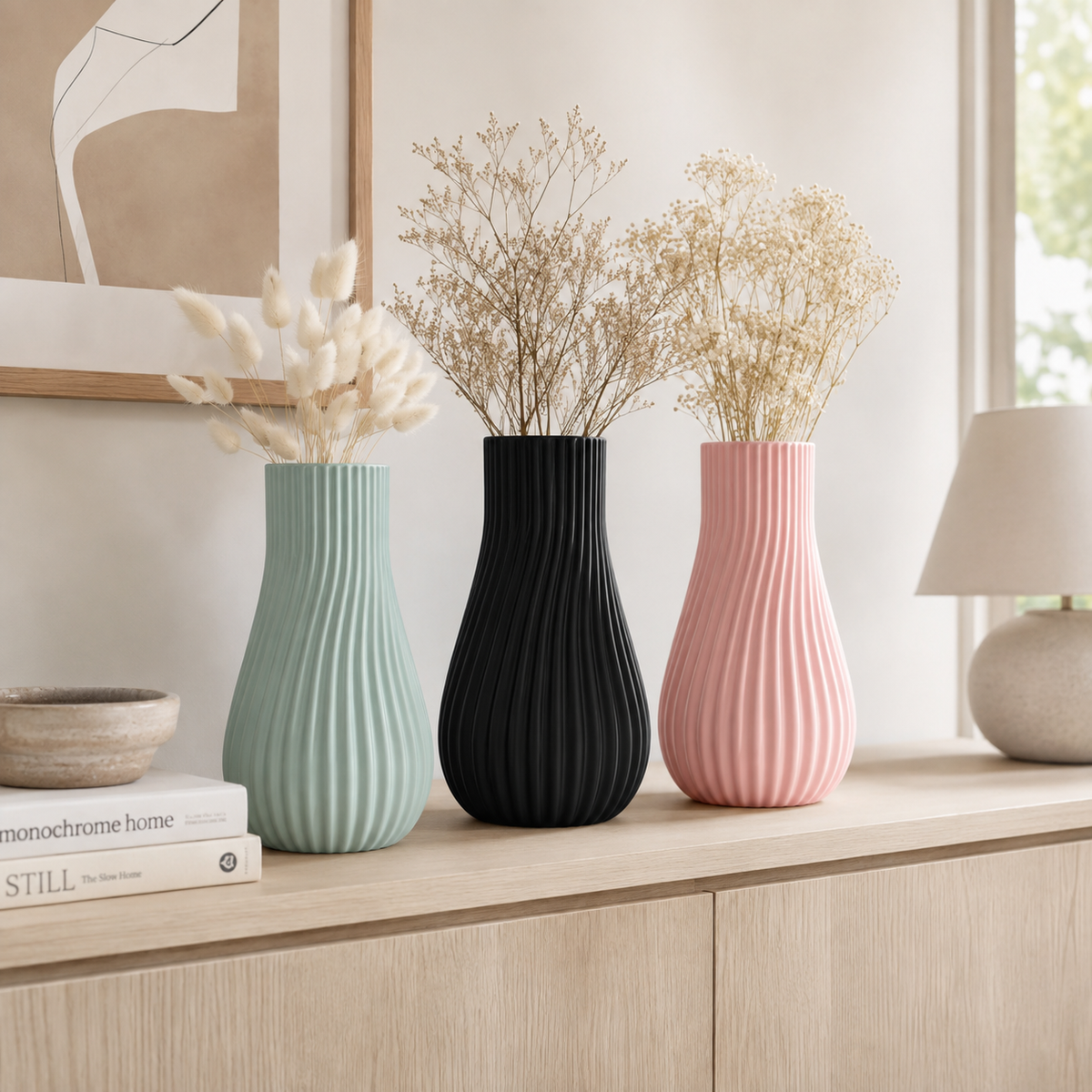 Three ribbed ceramic vases in mint green, black, and dusty pink, each filled with dried white flowers, arranged on a light wood console table in a bright, modern room.