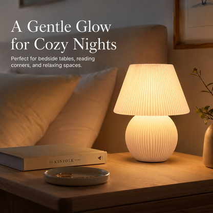 A warm white ribbed table lamp glowing on a wooden bedside table next to a book and small dish, with a beige headboard and pillows in the background. Text on the left reads: 'A Gentle Glow for Cozy Nights'.