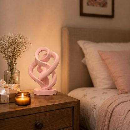 A pink sculptural table lamp with an abstract intertwined loop design sits on a wooden bedside table next to a lit candle, a glass vase with dried flowers, and a bed in a warmly lit bedroom.