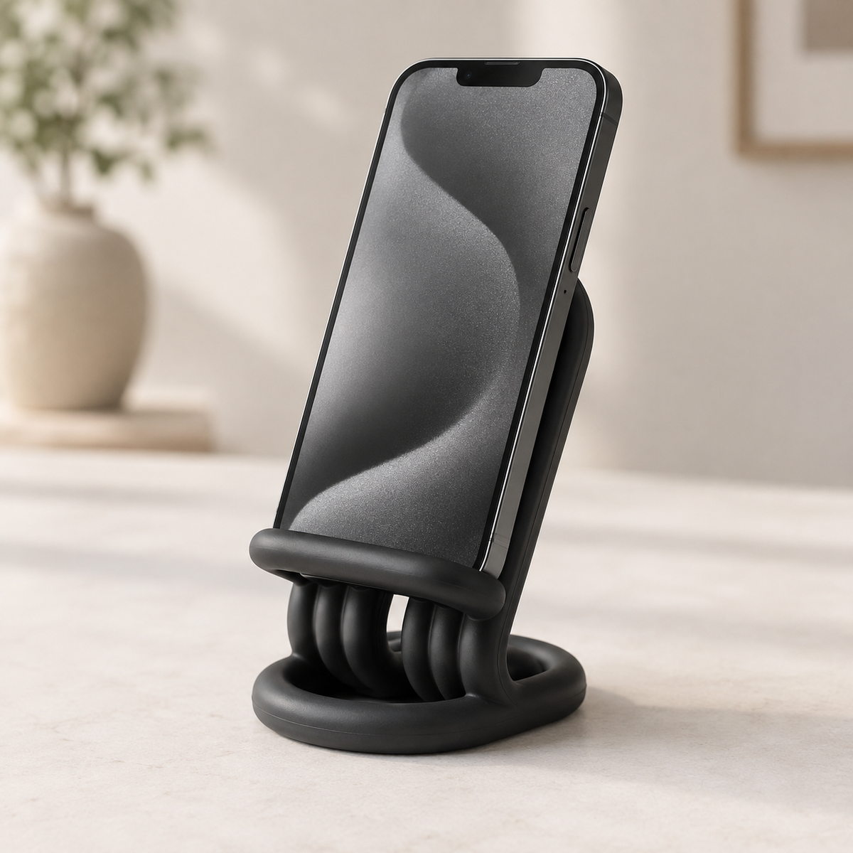A black silicone phone stand with a textured, ribbed base and a curved cradle holding a smartphone, set on a light beige tabletop with a blurred background.