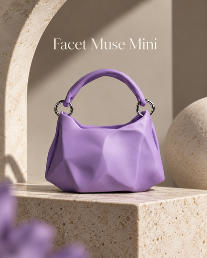 A small lavender purple handbag with a sculptural faceted design and a top handle, displayed on a stone pedestal in a minimalist architectural setting. The text 'Facet Muse Mini' is visible at the top.