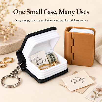 A small keychain ring box shown open and closed on a beige styled surface. The open case has a black exterior, white interior, and holds rings and folded cash; the closed case beside it is tan/brown.