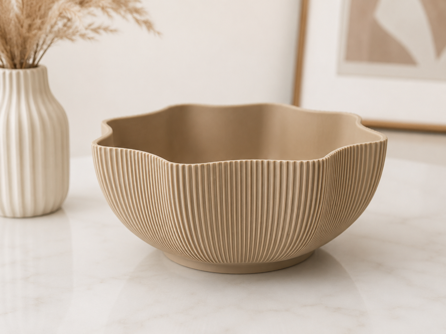 A beige ribbed ceramic bowl with a scalloped rim sits on a white marble surface next to a matching ribbed vase with dried grass stems.