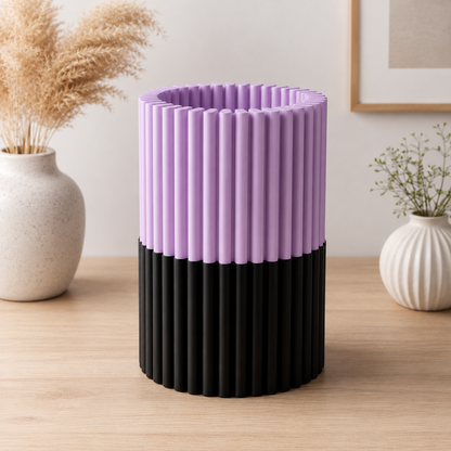 A ribbed cylindrical vase or planter with a two-tone design, featuring a lavender purple top half and a matte black bottom half, displayed on a light wood table with dried pampas grass and small plants in the background.