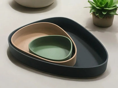 Three nested organic-shaped ceramic dishes in dark charcoal, beige, and sage green, arranged on a light beige surface with a small potted succulent in the background.