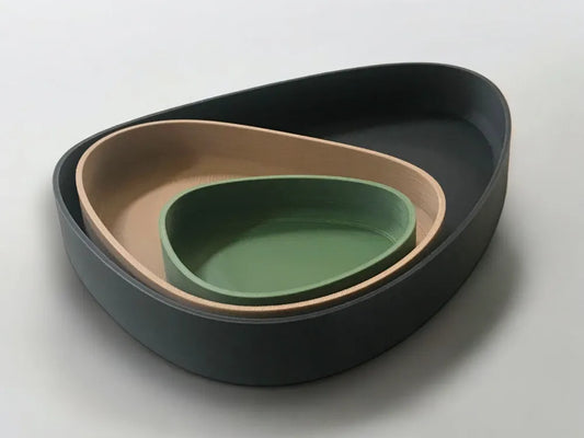 Three nested oval ceramic bowls in matte black, warm beige, and sage green, arranged on a light gray background.