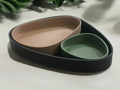 A three-piece matte ceramic tray set in black, beige, and sage green, arranged on a light surface with soft shadows and blurred green leaves in the background.
