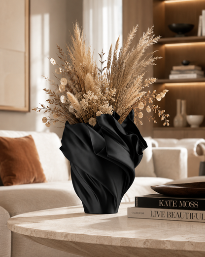 A black draped satin vase filled with dried beige pampas grass and decorative botanicals, placed on a light stone round table in a softly lit living room. Books and a sofa are visible in the background.