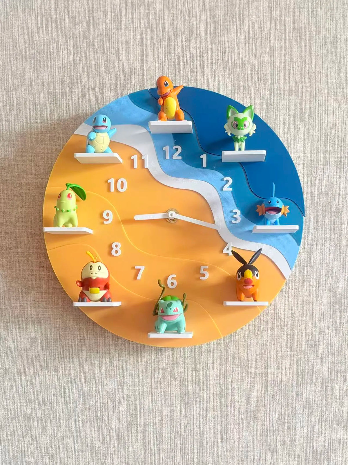 A circular wall clock featuring colorful Pokémon characters placed on white shelf-like platforms instead of traditional hour markers. The clock face is split into blue and orange sections with a white winding river-like divider, and the hands are white.