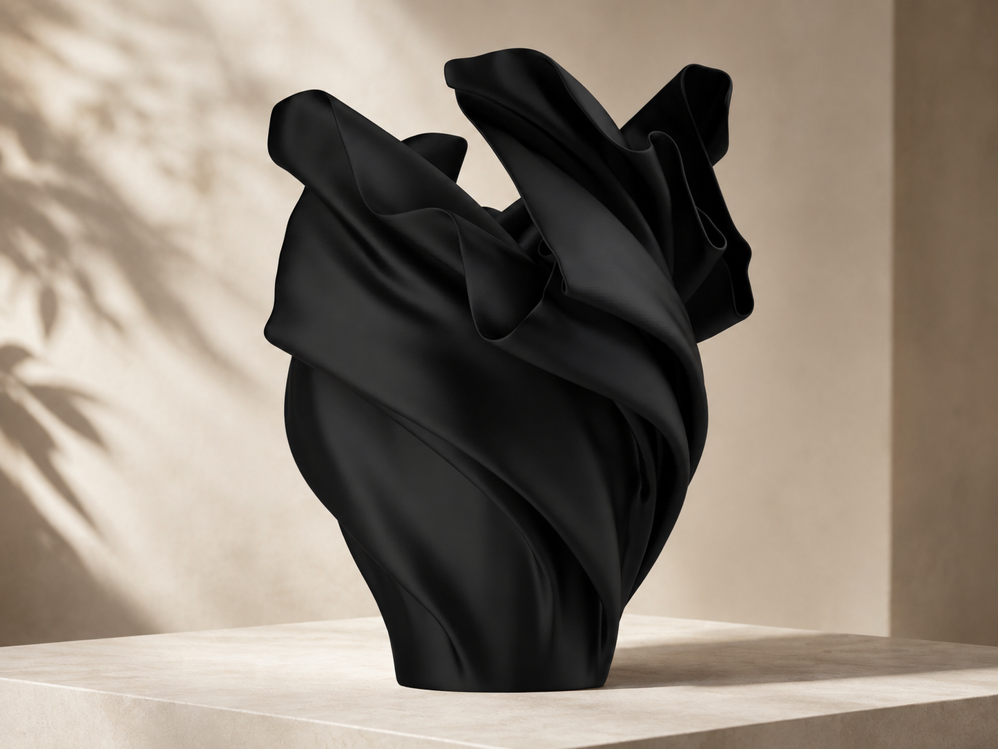 A sculptural black vase with a draped, flowing silhouette, displayed on a light beige stone pedestal against a warm beige wall with soft leaf shadows.