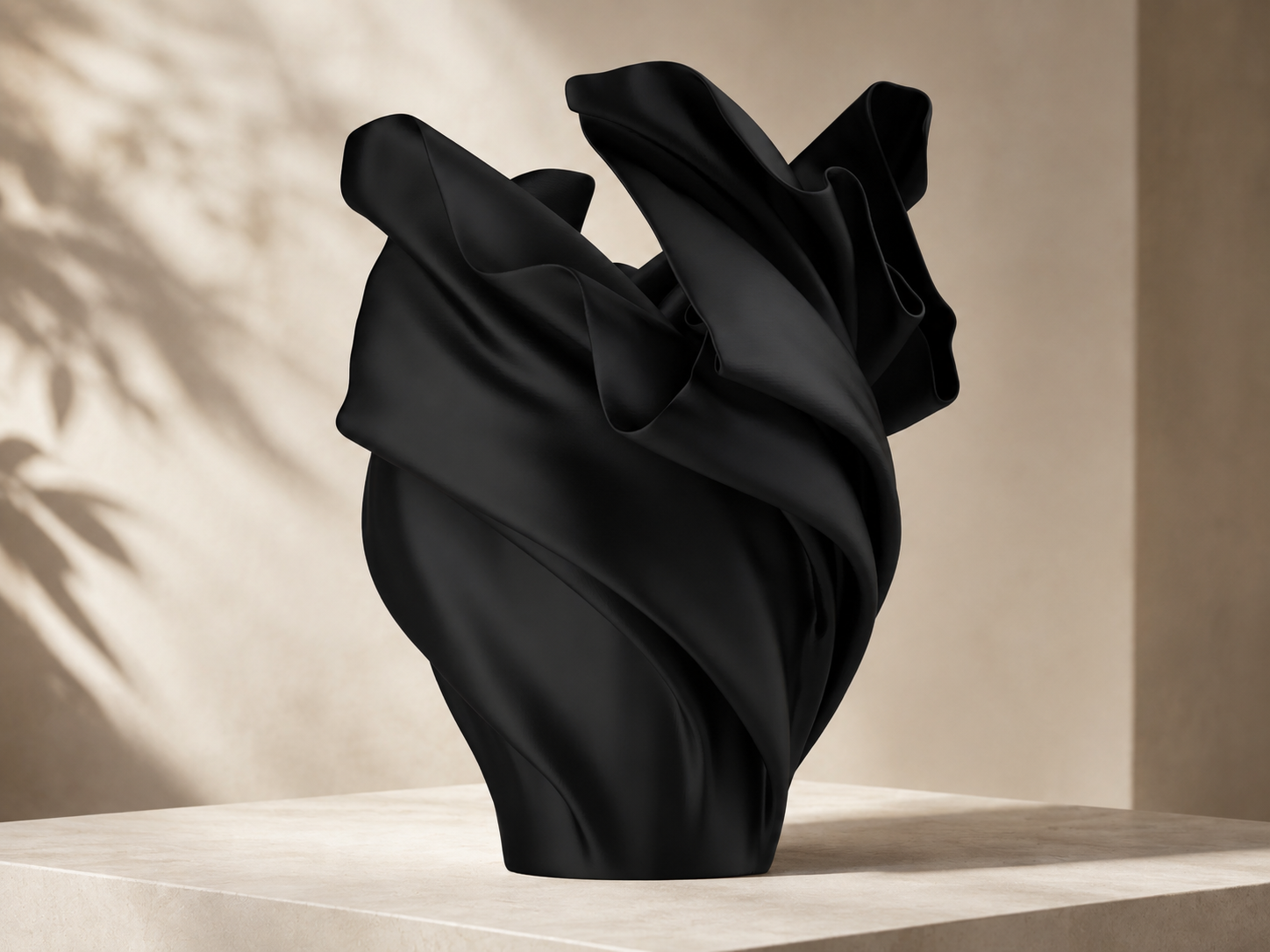 A sculptural black vase with a draped, flowing silhouette, displayed on a light beige stone pedestal against a warm beige wall with soft leaf shadows.