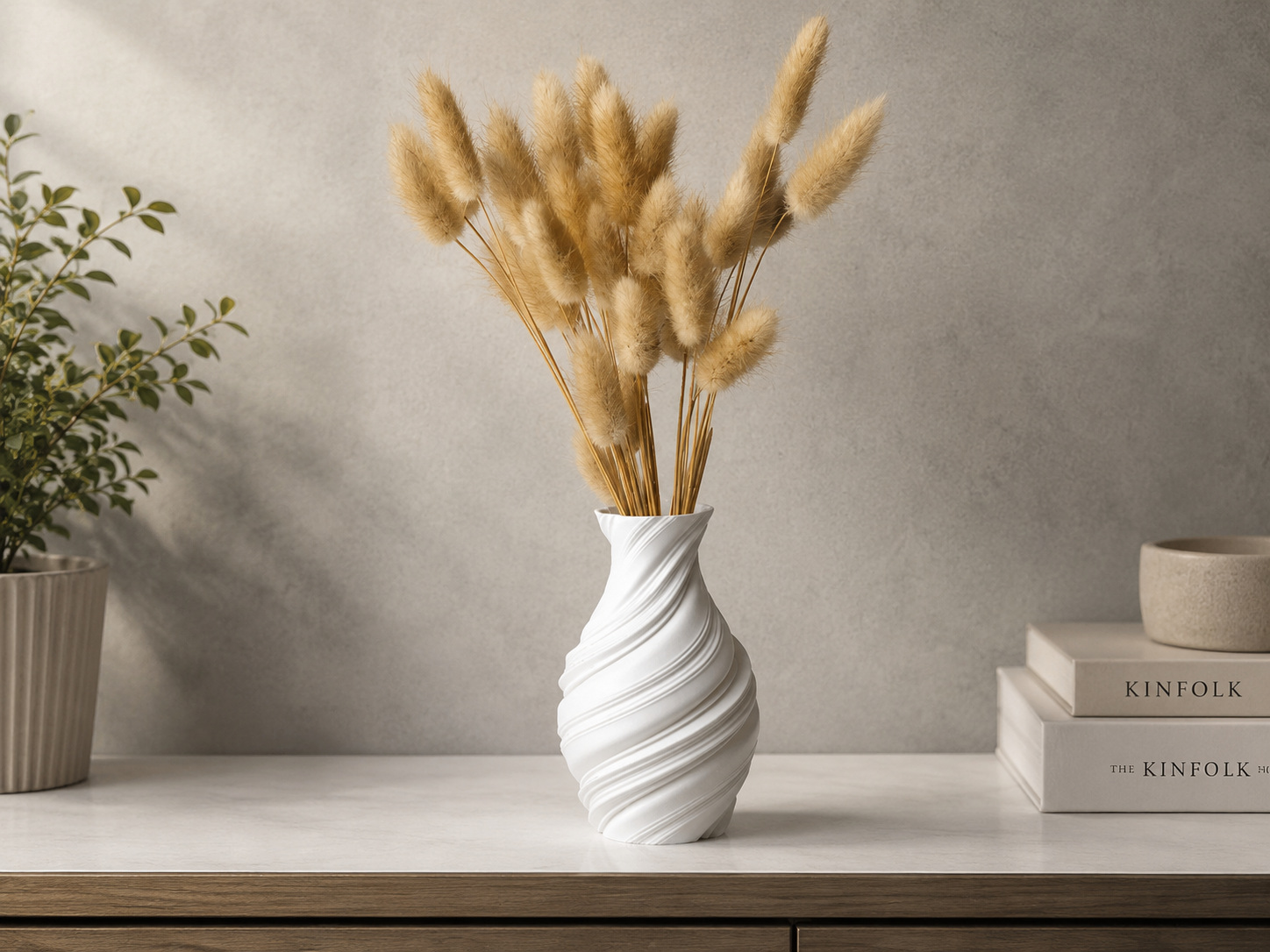 A white spiral-patterned ceramic vase holding dried pampas grass, styled on a light wood console table against a textured gray wall, with decorative books and a potted plant in the background.