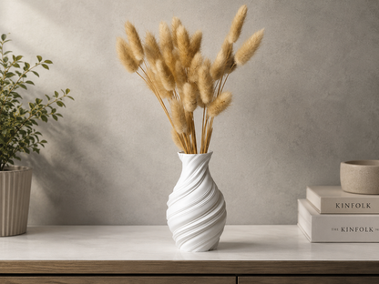 A white spiral-patterned ceramic vase holding dried pampas grass, styled on a light wood console table against a textured gray wall, with decorative books and a potted plant in the background.