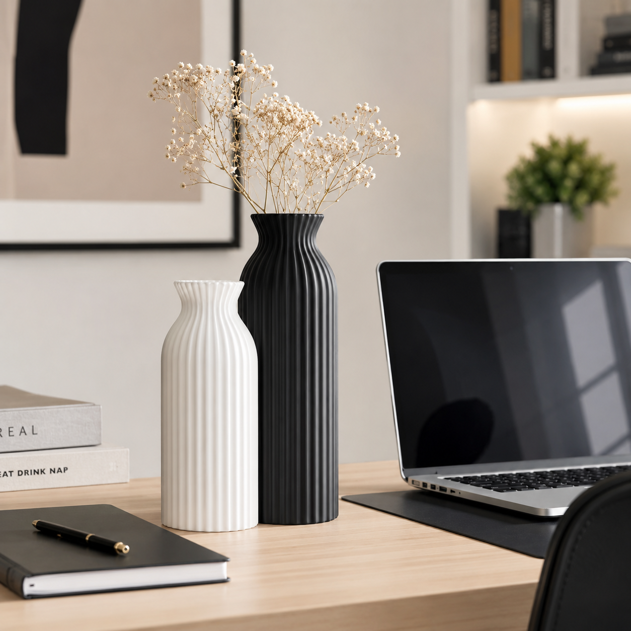 Two ribbed ceramic vases, one white and one black, with dried baby's breath stems, styled on a wooden desk next to a laptop in a modern home office setting.
