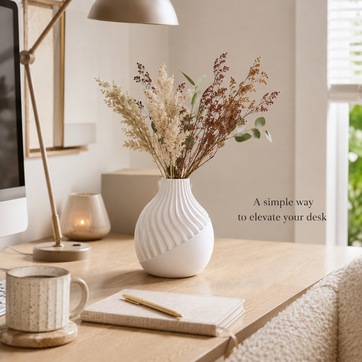 A white ribbed ceramic vase filled with dried flowers and greenery, styled on a wooden desk beside a mug, notebook, lamp, and candle in a bright home office setting.