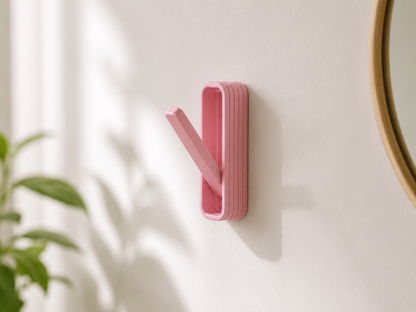 A pink wall-mounted hook with a slanted pull tab, shown mounted on a white wall with soft shadows and a blurred plant in the foreground.