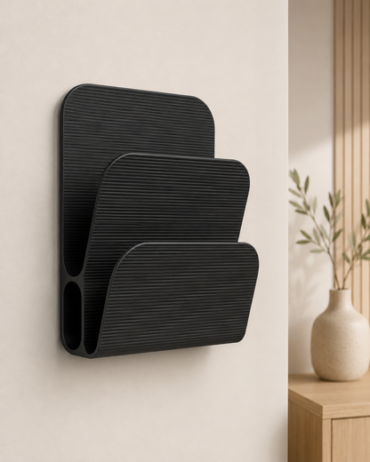 A black wall-mounted organizer with three tiered, ribbed compartments, shown mounted on a light beige wall in a modern interior setting with a wooden side table and a vase in the background.