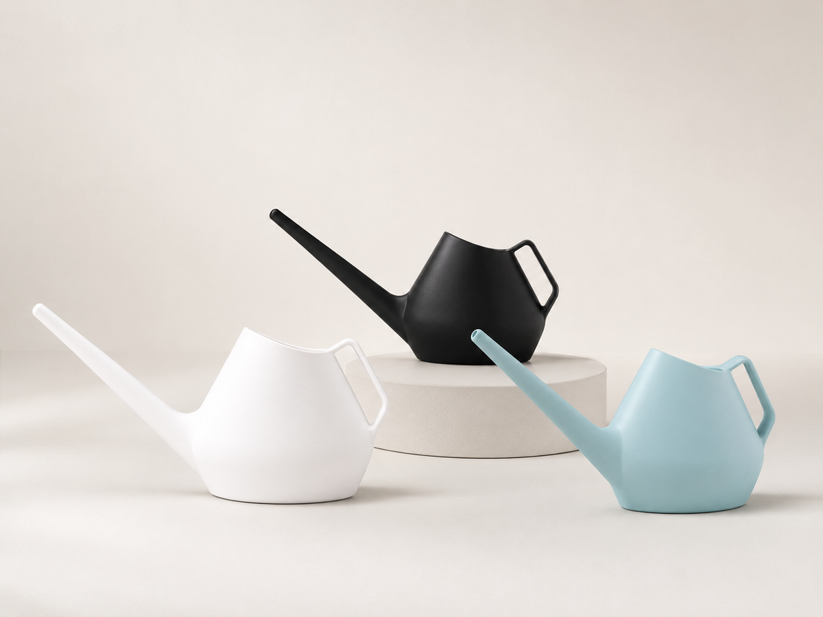 Three matte plastic watering cans in white, black, and light blue, arranged on a light beige background with a small round pedestal.