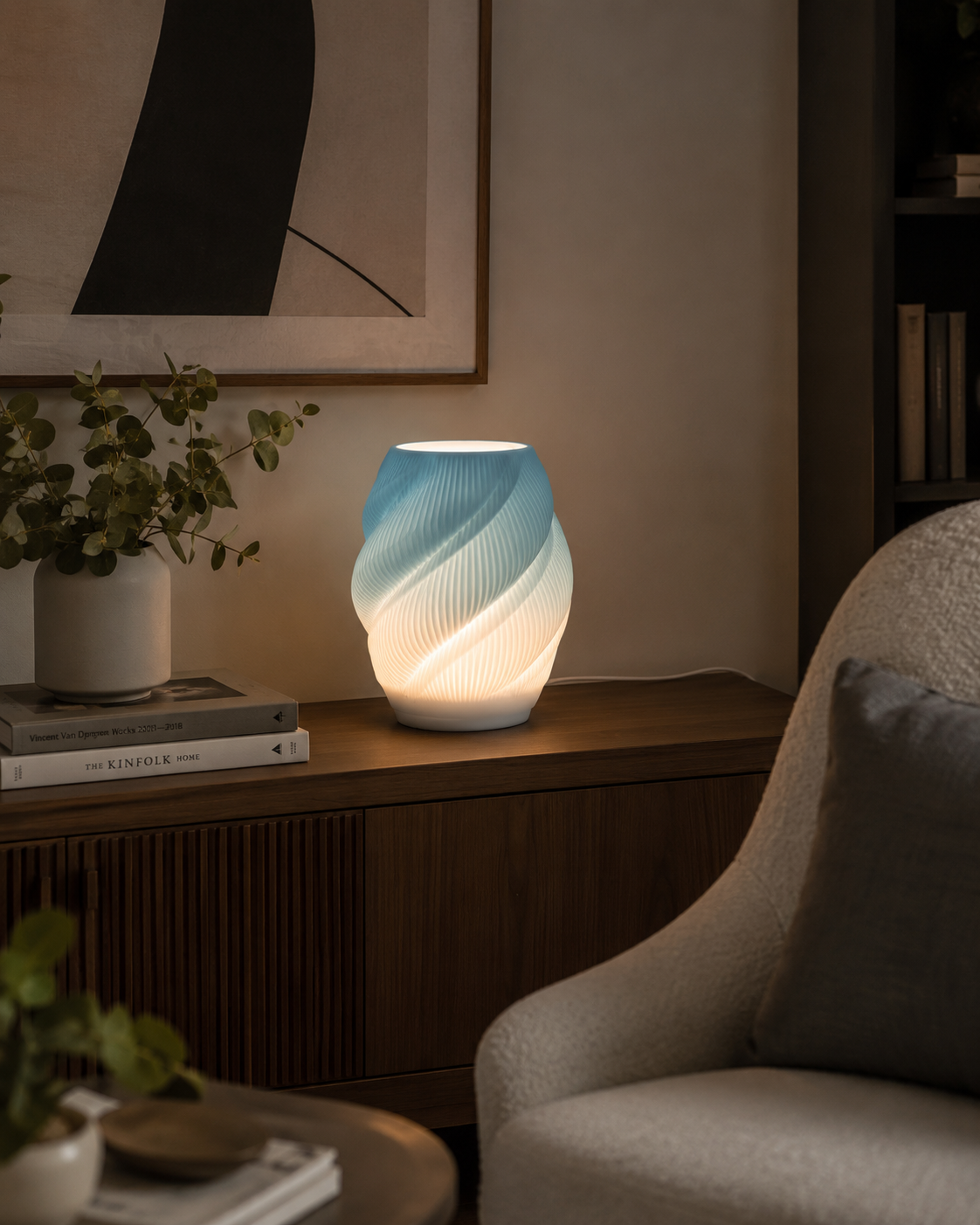 A modern spiral-shaped table lamp glowing with a blue-to-white gradient light, styled on a wooden sideboard in a cozy living room setting next to a chair.