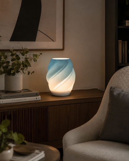A modern spiral-shaped table lamp glowing with a blue-to-white gradient light, styled on a wooden sideboard in a cozy living room setting next to a chair.